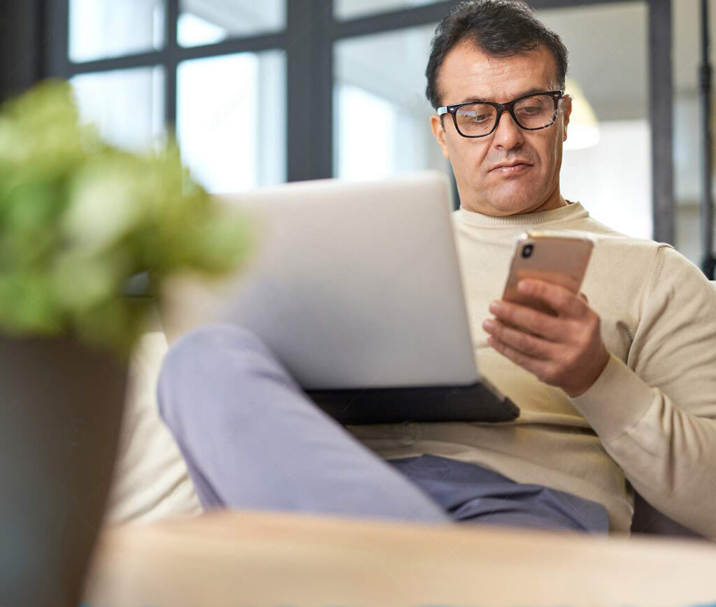 Un hombre utilizando un smartphone y un portátil, posiblemente gestionando inversiones en línea.